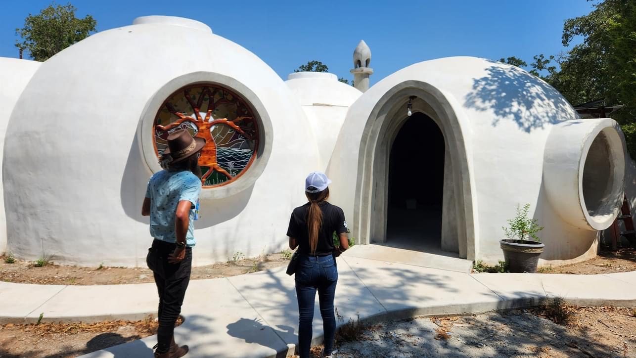 Two people stand before a completed white aircrete dome with a round stained-glass tree-of-life window and an arched doorway.