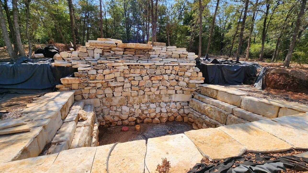Pool excavation with stacked limestone walls and steps under construction among cedar trees.